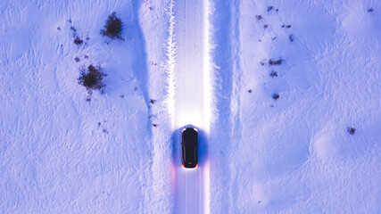 Aerial top view of black car driving on winter road in rural area while headlights illuminate in the dark, bird's eye view of suv vehicle crossing snowy cold lands with insurance. Lighting the way © BullRun