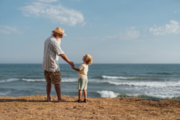 Grandfather and grandson standing together by the ocean, holding hands and admiring the waves,...