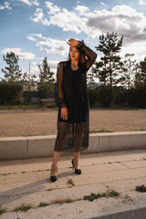 A stylish woman wearing a sheer black outfit and high heels poses gracefully under the sunlight in a serene outdoor setting.