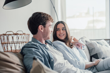 Young happy couple having fun talking laughing relaxing at home on couch, boyfriend embracing...