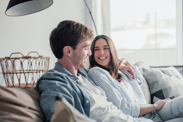 Young happy couple having fun talking laughing relaxing at home on couch, boyfriend embracing...