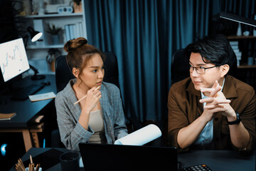 Two managers with stressful face discussing document paperwork file together comparing with report on laptop at over late night time at modern office, working on startup company project job. Infobahn.