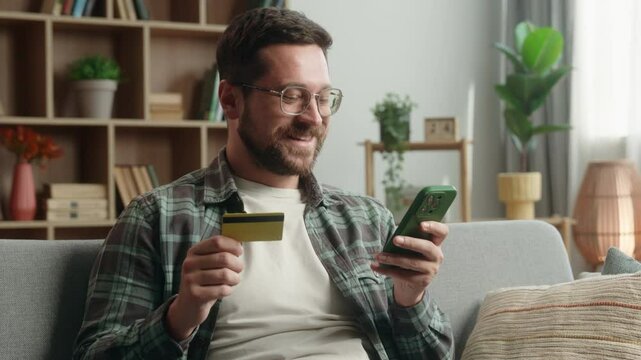 Smiling satisfied Caucasian adult addict man male guy shopping mobile phone at home couch payment online buying internet purchase holding credit card web money cashless e-commerce buy smartphone app