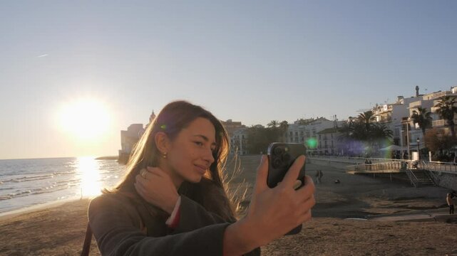 Beautiful young woman taking selfies, stories, and filming reels for social media with sea in background. Video call with friends through a mobile app. Sunset in Sitges