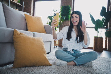 Happy young woman video calling while sitting on floor in bright cozy living room