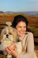 Cheerful laughing woman hugging her shih tzu dog in sunny weather against blue sky (part of the 