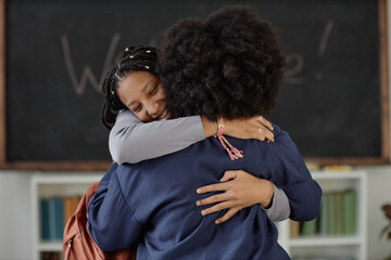 Waist up shot of teenage African American girl embracing teacher in classroom and smiling happily...