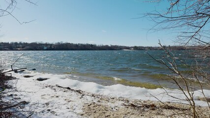 A peaceful winter scene at Lake Ronkonkoma, Long Island. Clear blue skies reflect over shimmering icy waters, creating a tranquil moment of natural beauty. Captures the serene charm of the season.