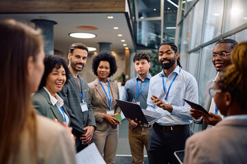 Black entrepreneur using laptop while talking to group of business seminar attendees in hallway.