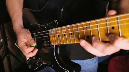 Fototapeta premium A close-up shot of a person playing a black electric guitar. The focus is on the hands, one holding a green pick and the other pressing the strings on the fretboard.