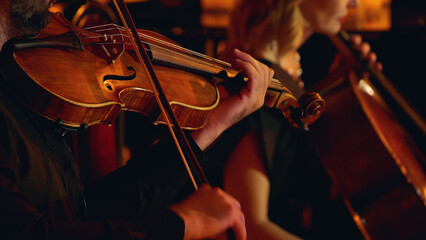 A close-up of musicians playing string instruments in an orchestra, focusing on a violinist and a cellist. The warm lighting highlights the wood of the instruments.