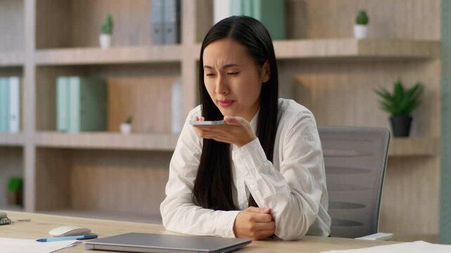 Irritated annoyed unhappy boss Japanese office worker business woman ethnic Chinese Korean female businesswoman girl using smartphone recording voice message send audio SMS talking loudspeaker indoors