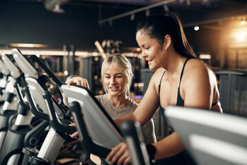 Smiling female instructor offering encouragement to a fit young woman riding on a stationary bike during a cardio exercise session at the gym