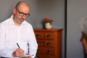 A therapist listens attentively and takes notes during a confidential meeting with a mature female client in a modern office.