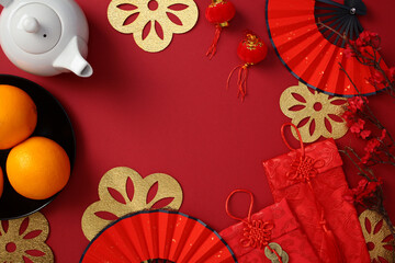 Obraz premium Chinese New Year celebration background with traditional elements: red fans, gold decorations, oranges, teapot, red envelopes, and cherry blossoms on a red background