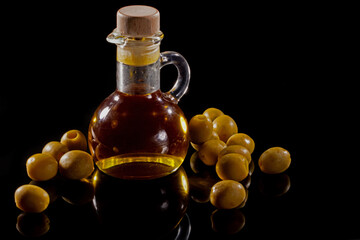 Glass jug with olive oil and scattered olives on black mirror background