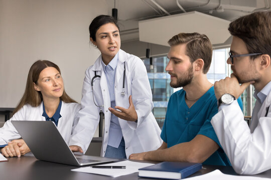 Group of medical professionals gathered in conference room with laptop, led by young Indian team leader explain, discuss topic, share treatment plan, offering solution, consider patient case together