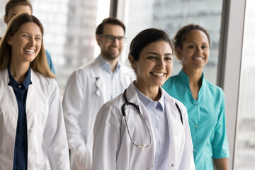 Group of diverse doctors, clinic staff posing together led by Indian woman in white lab coat, smile, look into distance with confident expression, exude leadership, professionalism in healthcare field