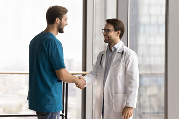 Two Caucasian men doctors standing in clinic, shaking hands, have reached consensus treatment plan of hospital patient, represent successful collaboration, completing challenging together or teamwork