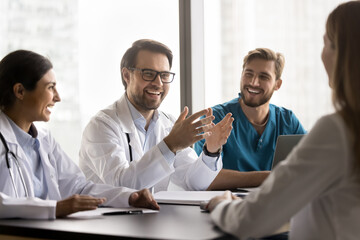 Laughing doctors mates gathered at conference table, lead talk, giggling, enjoy pleasant friendly collaboration in skyscraper clinic office, medical briefing led by young Caucasian man chief. Medicine