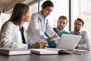 Obraz premium Indian GP professional share experience, opinion, treatment plan regarding patient diagnose results. Group of young skilled medical workers in uniforms engaged in teamwork, collaboration using laptop