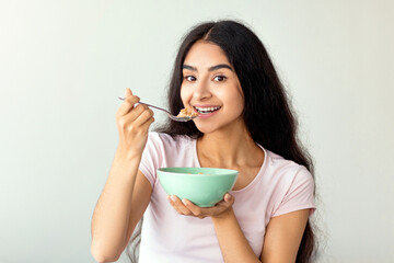 Attractive Indian lady having cereal with milk or cornflakes while sitting on comfy bed at home,...