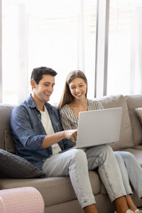 Positive young Hispanic couple using online application on laptop for media entertainment at home, enjoying modern Internet technology, shopping on ecommerce platform together. Vertical shot