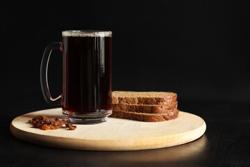 Mug of tasty kvass, bread and raisins on a dark background. Glass mug of kvass on a wooden board, close-up. Dark kvass with ingredients on a black background. Refreshing drink with slices of bread
