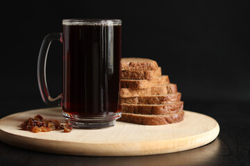 Mug of tasty kvass, bread and raisins on a dark background. Glass mug of kvass on a wooden board, close-up. Dark kvass with ingredients on a black background. Refreshing drink with slices of bread