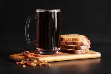 Mug of tasty kvass, bread and raisins on a dark background. Glass mug of kvass on a wooden board, close-up. Dark kvass with ingredients on a black background. Refreshing drink with slices of bread