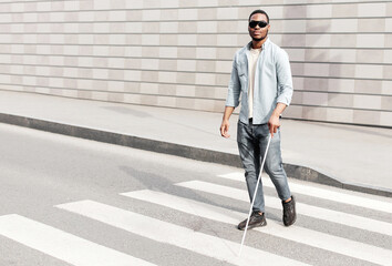 Full length of young black man with visual disability wearing dark glasses, using walking cane to...