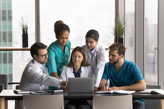 Five multiethnic clinic staff members, professional medical workers working together using laptop, review latest medical research, treatment protocols related to patient condition. Team collaboration