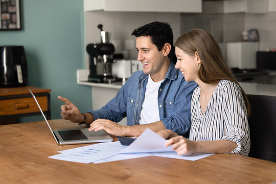 Cheerful young couple of new home owners paying paper bill, invoices at laptop, making online payment, virtual transaction, reviewing paper reports at kitchen table, smiling, talking
