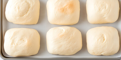 Six square-shaped bread dough pieces arranged on a baking tray, ready for baking. Soft, pale yellow dough awaits oven time.
