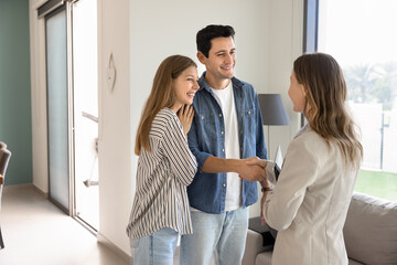 Happy young couple of buyers or renters shaking hands with real estate insurance agent, standing...