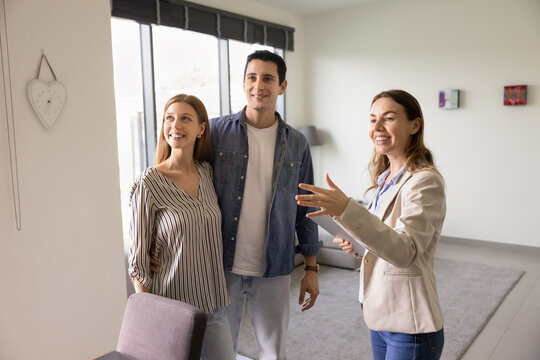 Happy realtor woman showing apartment to positive young couple of renters. Real estate agent and buyers meeting in house or flat, looking at interior, discussing buying, mortgage