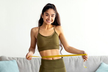 Full length of cheerful Hindu lady in sportswear standing on scales, measuring waist with tape, feeling happy about losing weight at home. Healthy lifestyle, sports and slimming diet concept
