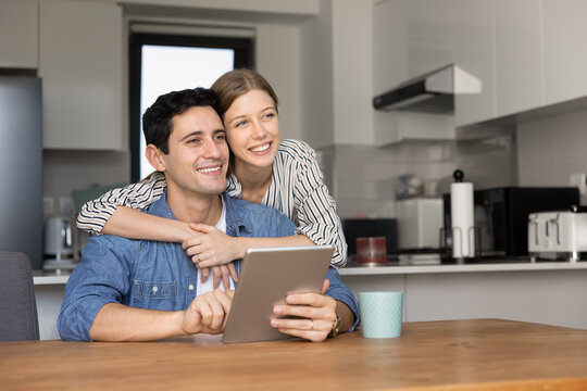 Happy dreamy young couple hugging in kitchen, using virtual assistant on digital tablet, looking away, thinking on modern smart home technology, holding gadget, smiling, laughing - Powered by Adobe