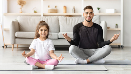 Sporty middle eastern father and kid sitting on fitness mat, doing yoga and smiling, home interior. Happy dad and daughter kid in sportswear having moring training together in living room