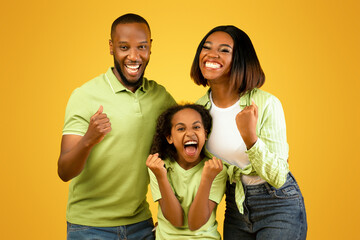 Victory concept. Emotional overjoyed black family of three shaking fists, gesturing yes and shouting, standing over yellow background. Young parents and daughter rejoicing success.