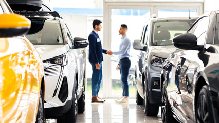 Middle-eastern young man customer and sales assistant shaking hands, car showroom interior, friendly manager greeting wealthy arab guy, standing among luxury brand new autos, full size shot