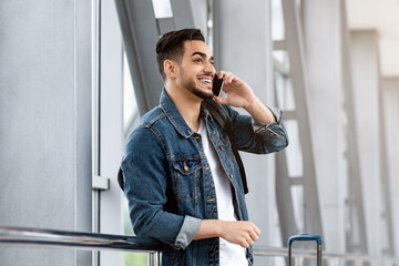 Mobile Communication. Smiling Young Arab Guy Talking On Cellphone At Airport, Millennial Middle Eastern Man Standing With Luggage Near Window At Terminal And Enjoying Pleasant Call, Copy Space