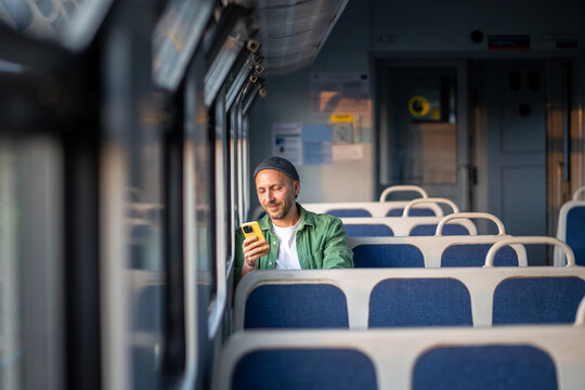 Smiling man using smartphone, scrolling social media and messaging, seated by the window in train in the morning. Public transit, morning travel, commute routine.