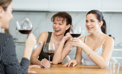 Merry middle-aged women are seated at a table drinking wine and talking in comfort of their house
