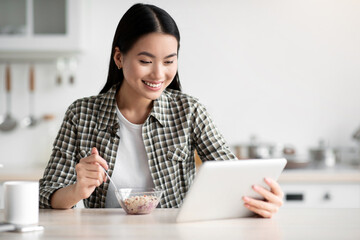 Positive asian woman having breakfast and surfing on social media, using digital tablet, kitchen...