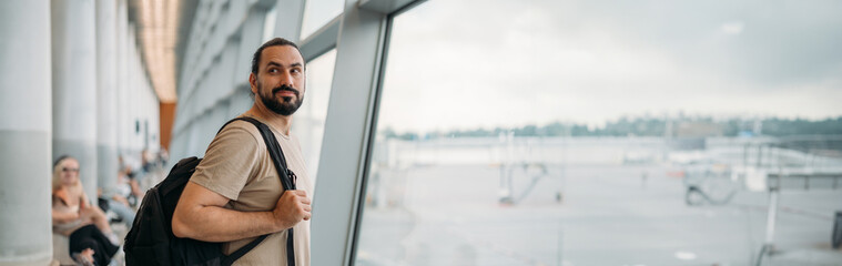 A young man with a backpack is waiting for departure at the airport. A Caucasian guy stands at a large window at the gate © Anna
