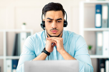 Worried millennial Arab man in headphones making online video call with business partner, having...