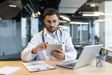 Obraz premium Focused doctor sitting at desk with laptop. Man in white medical coat consulting patients remotely at workplace in office.