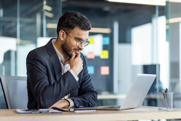 Thinking businessman at workplace inside office, man in business suit working with laptop, reading online report, and preparing presentation investment project.
