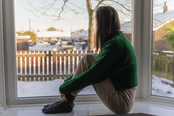 Young woman sitting by the window, looking out at a winter scenery outside. Coronavirus quarantine concept
